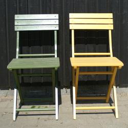 På billedet ser du to træstole udenfor. Stolene står foran en lodret træbeklædning som baggrund. Den venstre stol er malet i grønne nuancer og bærer spor af vejrpåvirkning, som afskallet maling og en lettere forvitret overflade. Den højre stol er malet i en ensartet gul farve og ser ny og velplejet ud. Begge stole har en simplistisk og funktionel design: et retvinklet sæde samt et lige ryglæn med horisontale planker. Farverne kan repræsentere udvalgte muligheder i vores sortiment af træbeskyttelsesmaling. Hver farve er tilgængelig i 5-liters spande.